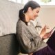 Woman wearing a gray sweatshirt sitting beside her bed and using her smartphone