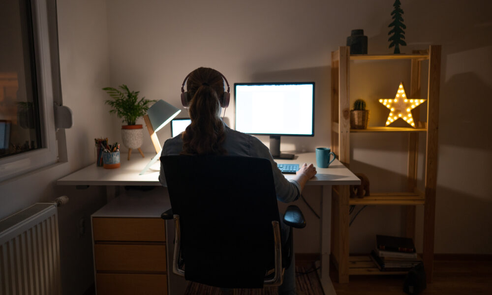 Bakifrån av en oigenkännlig ung kvinna som arbetar sent på sin dator från sitt hemmakontor.