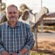 Peter wearing a blue check shirt, standing outside in the rural town of Norseman, with gumtrees and dirt.