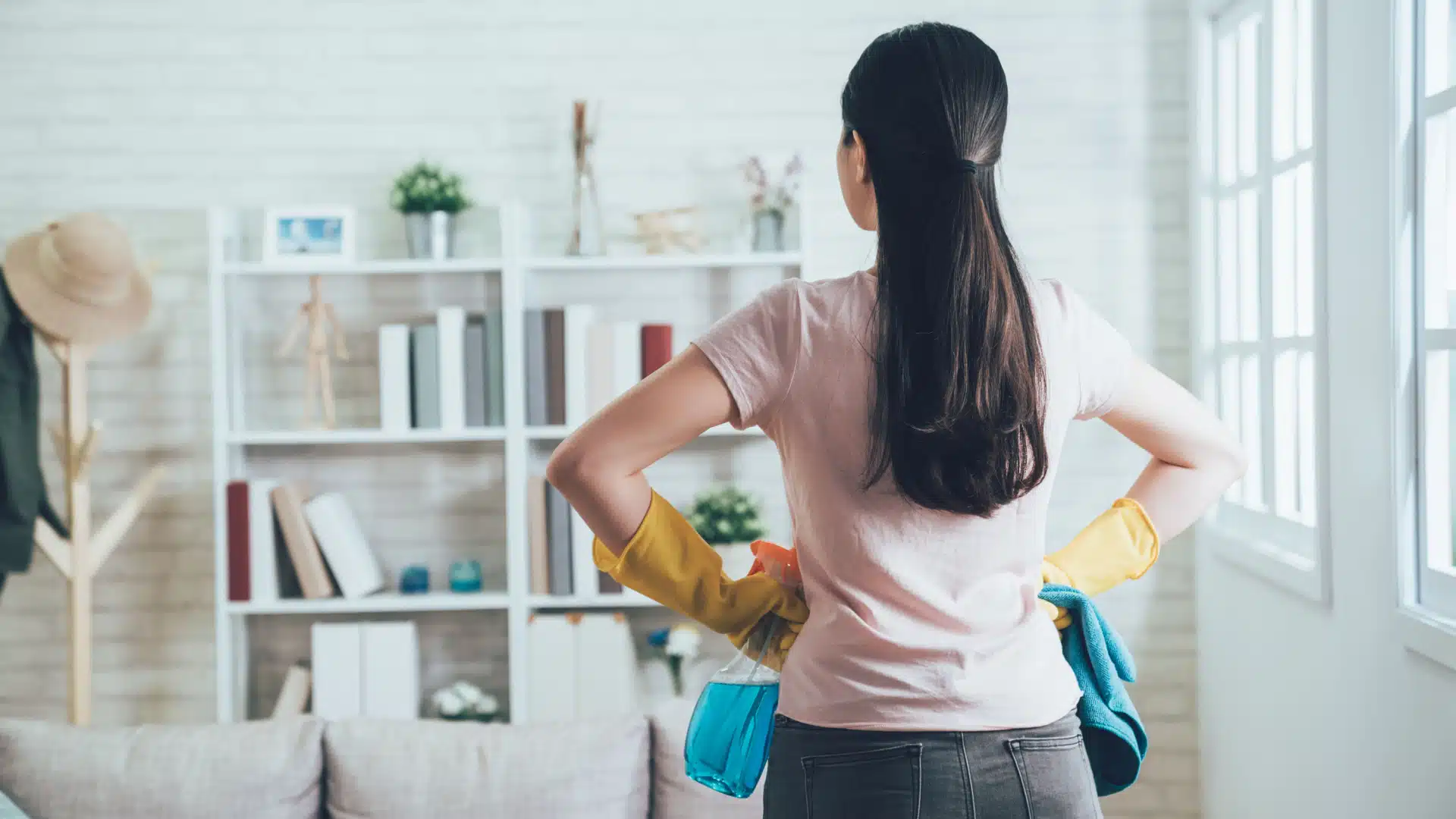 Woman looking away into a room with her back to the amera. She has her hands on her hips and cleaning supplies in them. She looks as if she's ready to tackle the chores.