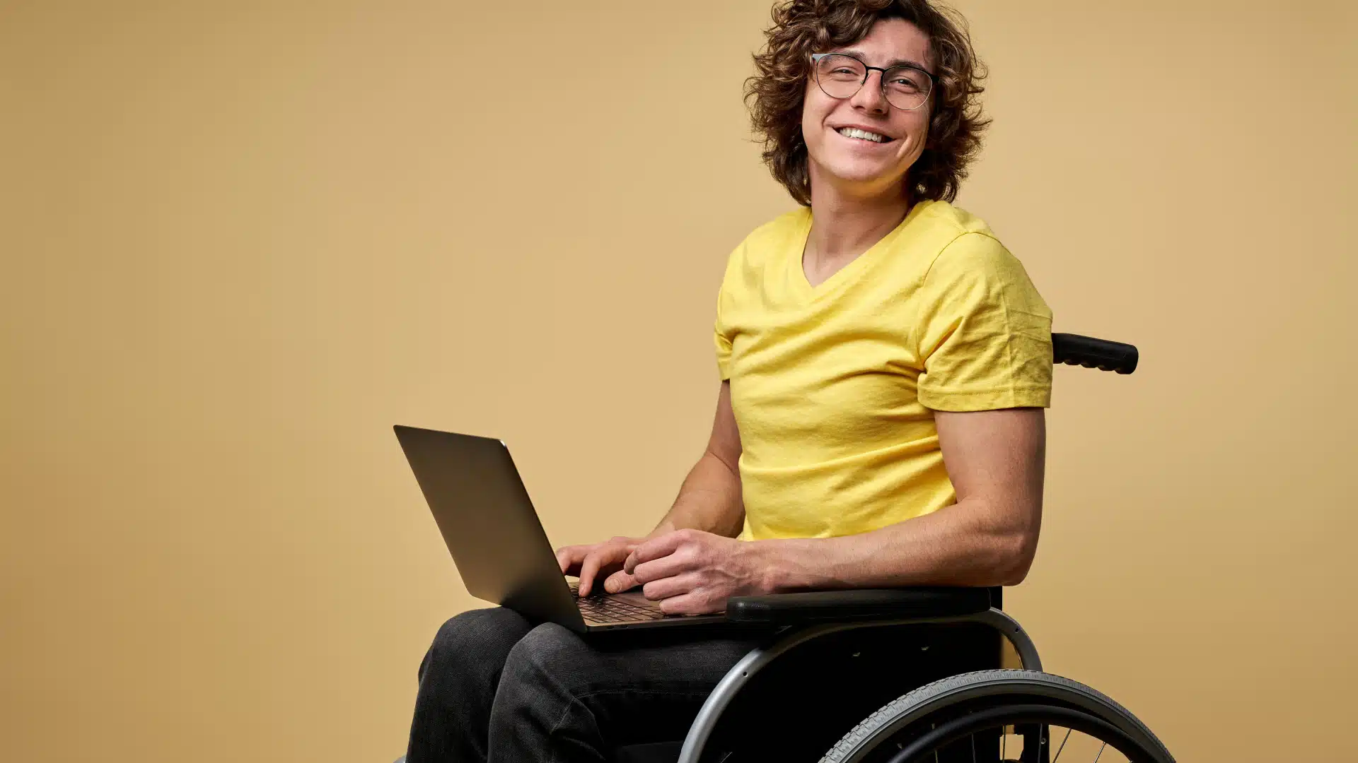 A man sits in a wheelchair with a laptop in his lap. He's turned towards the camera and smiling.
