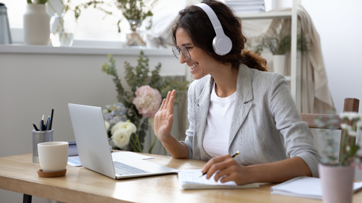 Smiling young Caucasian woman in headphones take online educational course or training on laptop from home, happy female in wireless headset wave to camera, talk on webcam video call on computer.