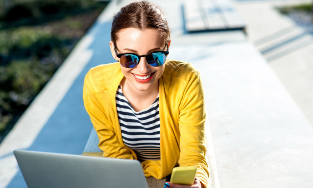 Young woman in yellow sweater working with laptop and phone on the bench in the city.