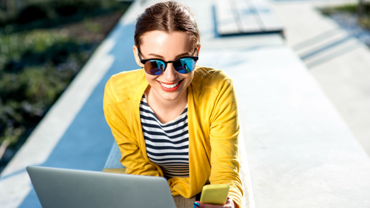Young woman in yellow sweater working with laptop and phone on the bench in the city.